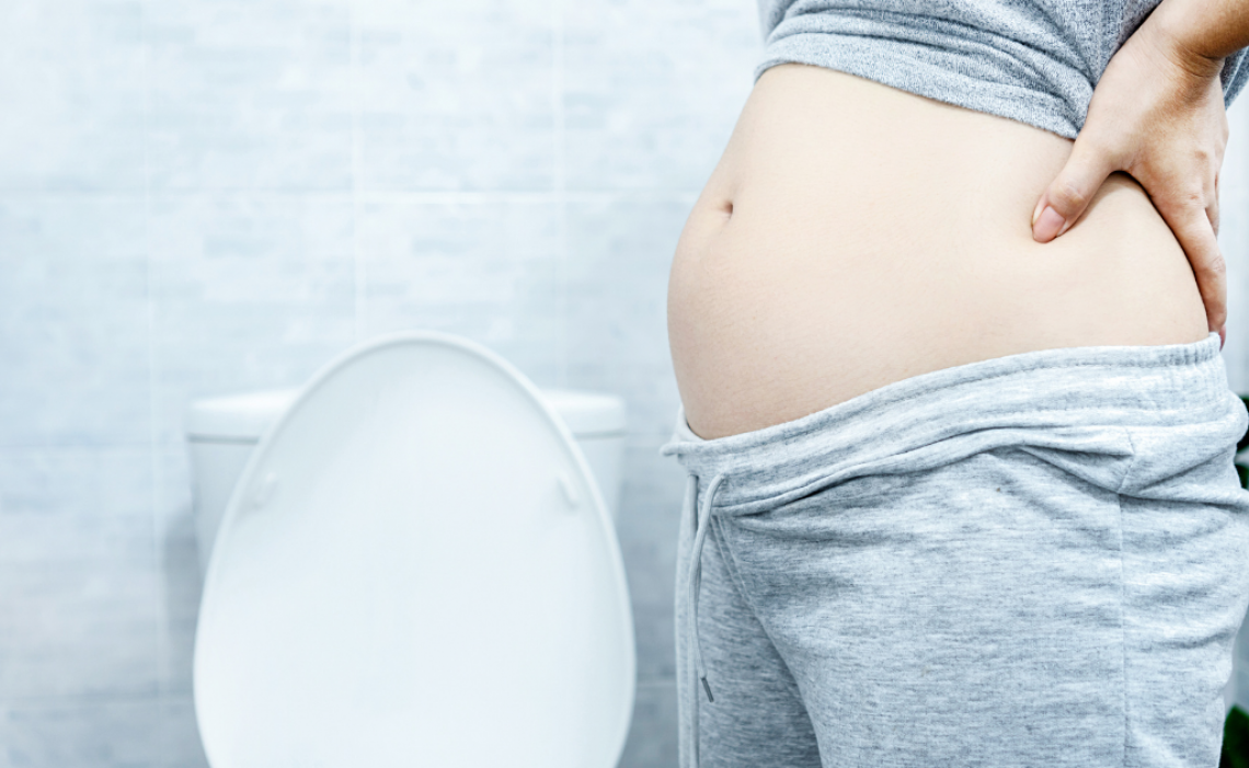 Frau steht mit schmerzhaft aufgeblähtem Bauch vor der Toilette – Symbolbild für Verdauungsbeschwerden, Blähbauch und Darmprobleme